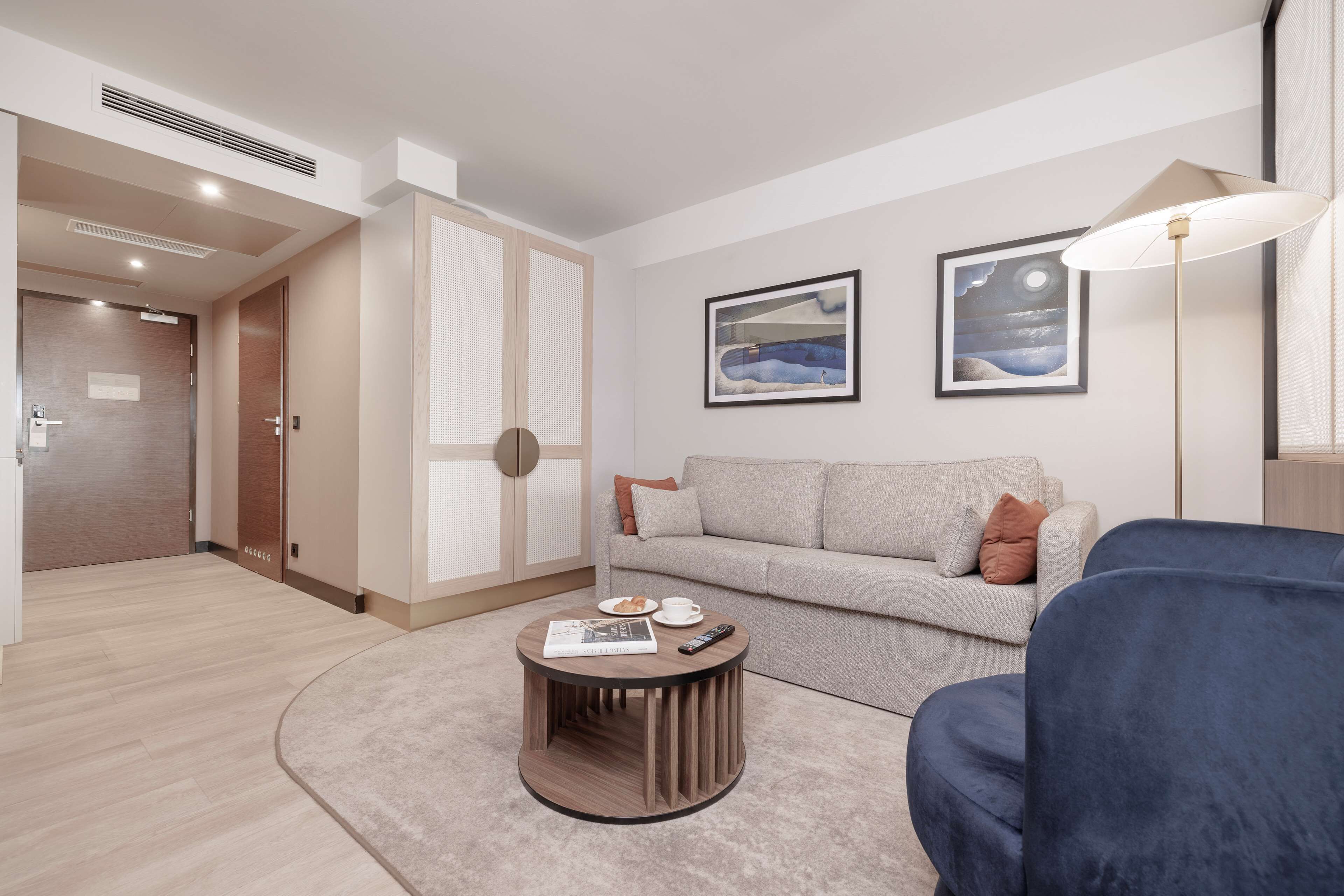 A modern living room with a round coffee table, a sofa, and a lamp. There are two framed pictures on the wall and a door leading to another room.