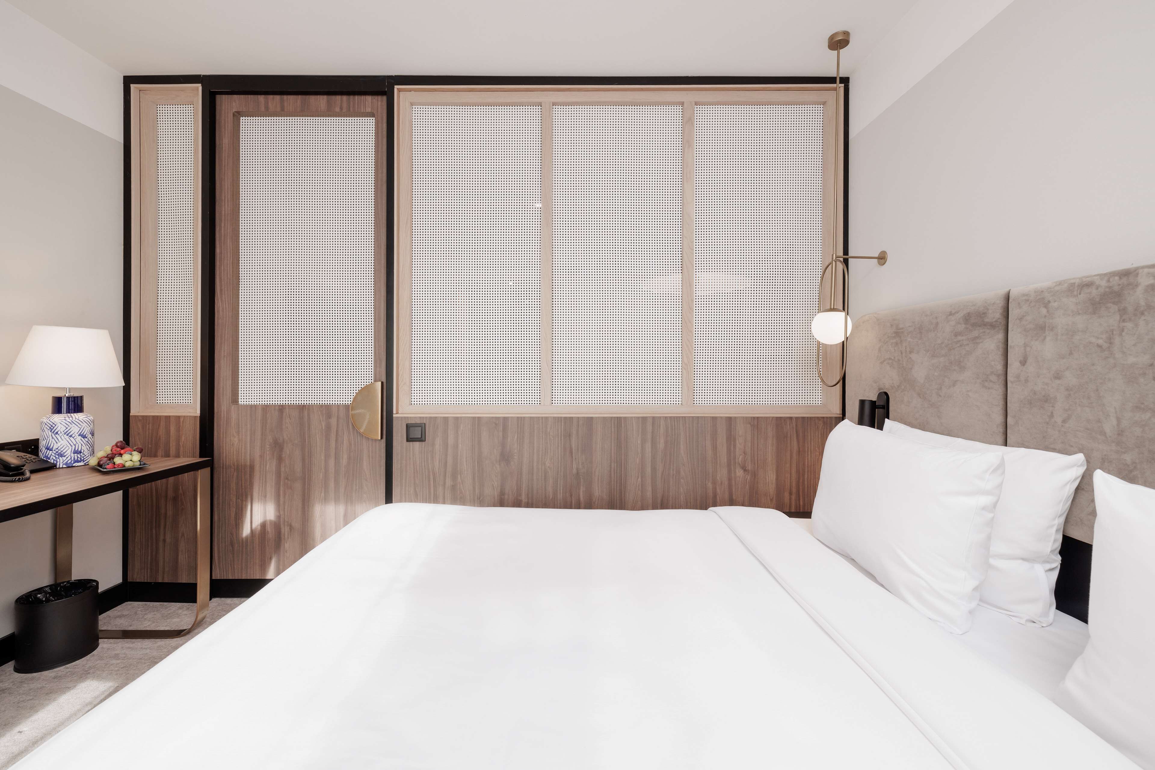 A hotel room with a large bed, a desk with a lamp, and a view through a wooden door with frosted glass panels.