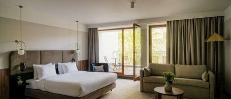 A hotel room with a large bed, a sofa, a coffee table with a plant, and a view of the outdoors through a sliding door.