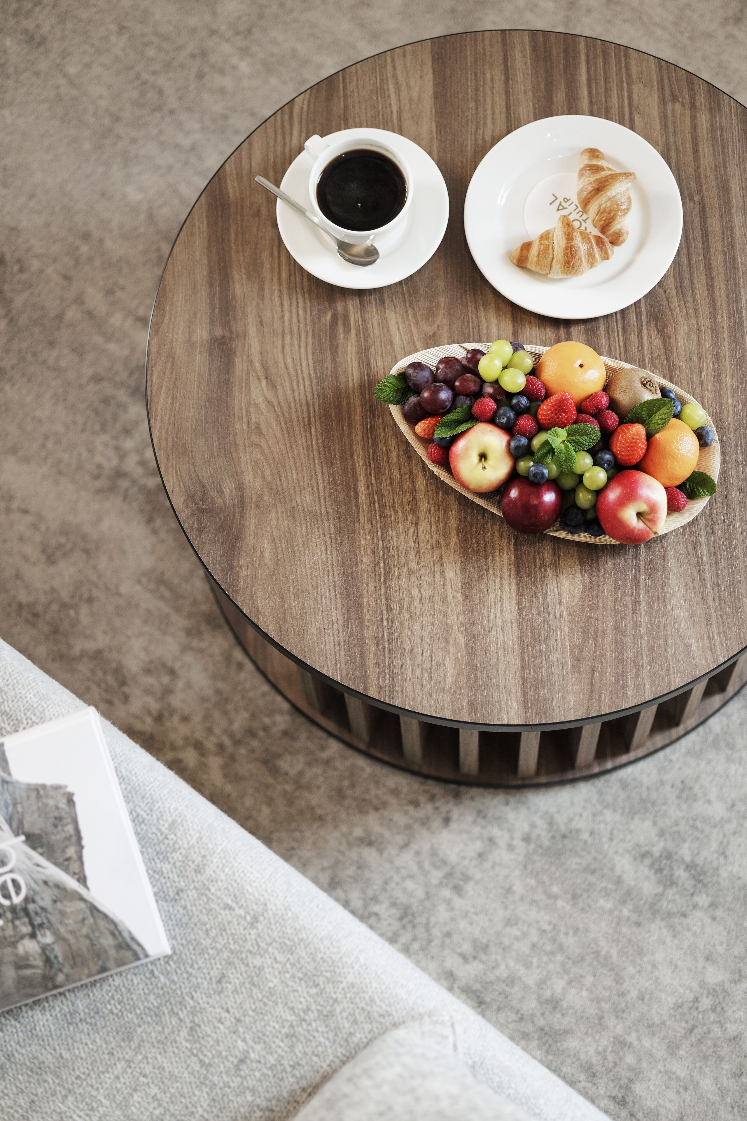 Una mesa de madera redonda con un plato blanco de croissants, una taza blanca de café y un bol de madera con frutas variadas.