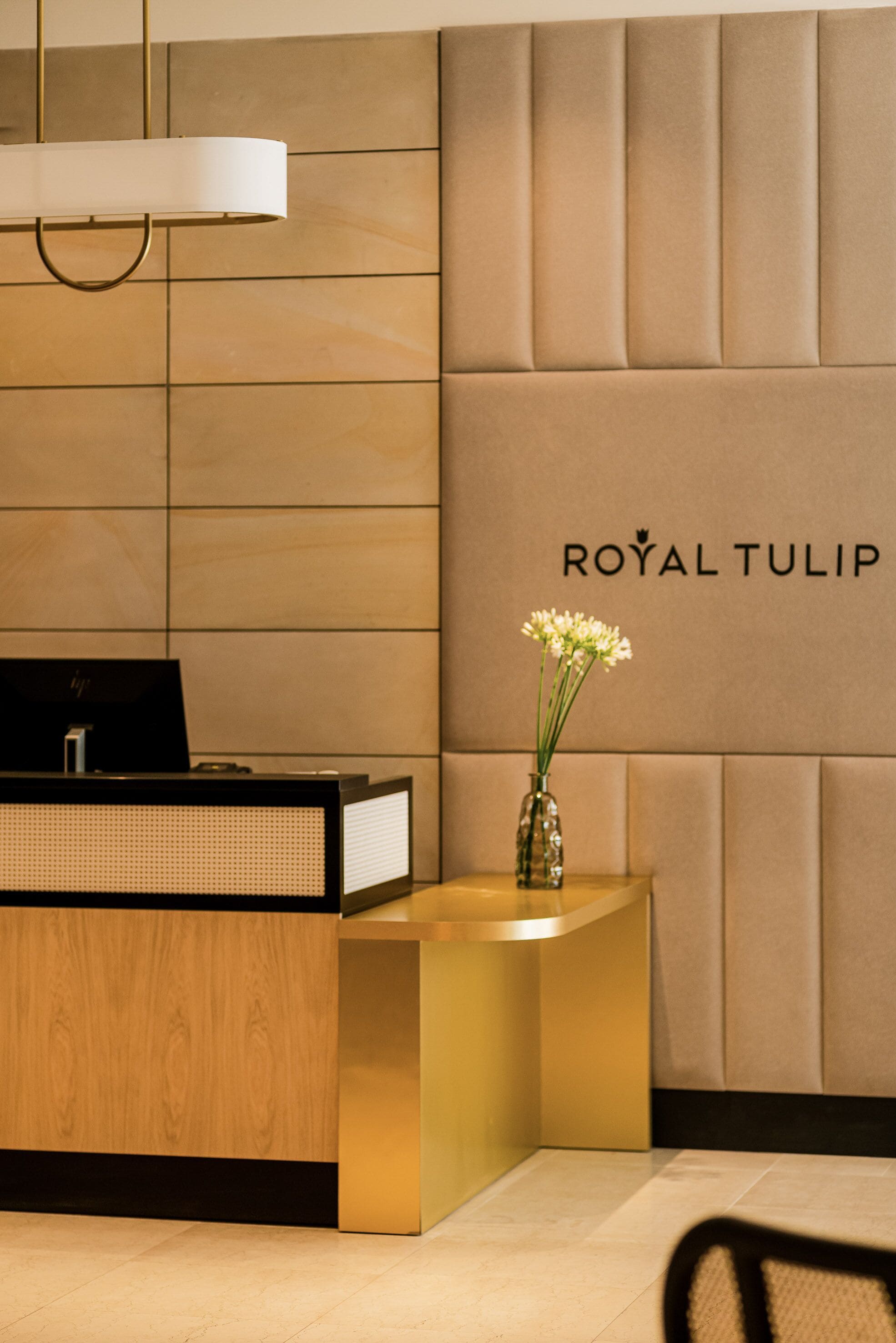 Ein Empfangsschalter mit Holzoptik, ein schwarzer HP-Monitor und eine Vase mit weißen Blumen. An der dahinterliegenden Wand ist eine gepolsterte Gestaltung zu sehen, und der Schriftzug "ROYAL TULIP" ist sichtbar.