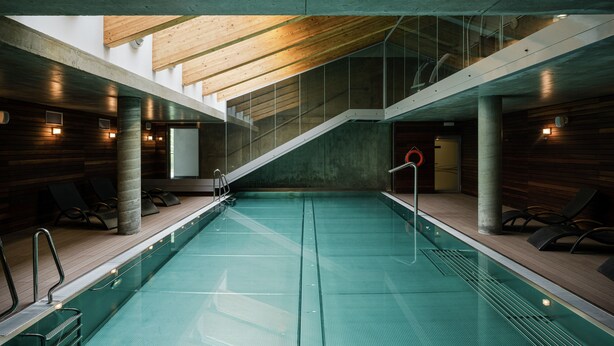 Una piscina interior moderna con techo y paredes de madera, barandillas de cristal transparente y sillones de descanso a los lados.