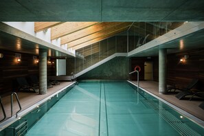 Ein moderner, überdachter Pool mit Holzdecke und -wänden, klaren Glasgeländern und Liegestühlen entlang der Seiten.
