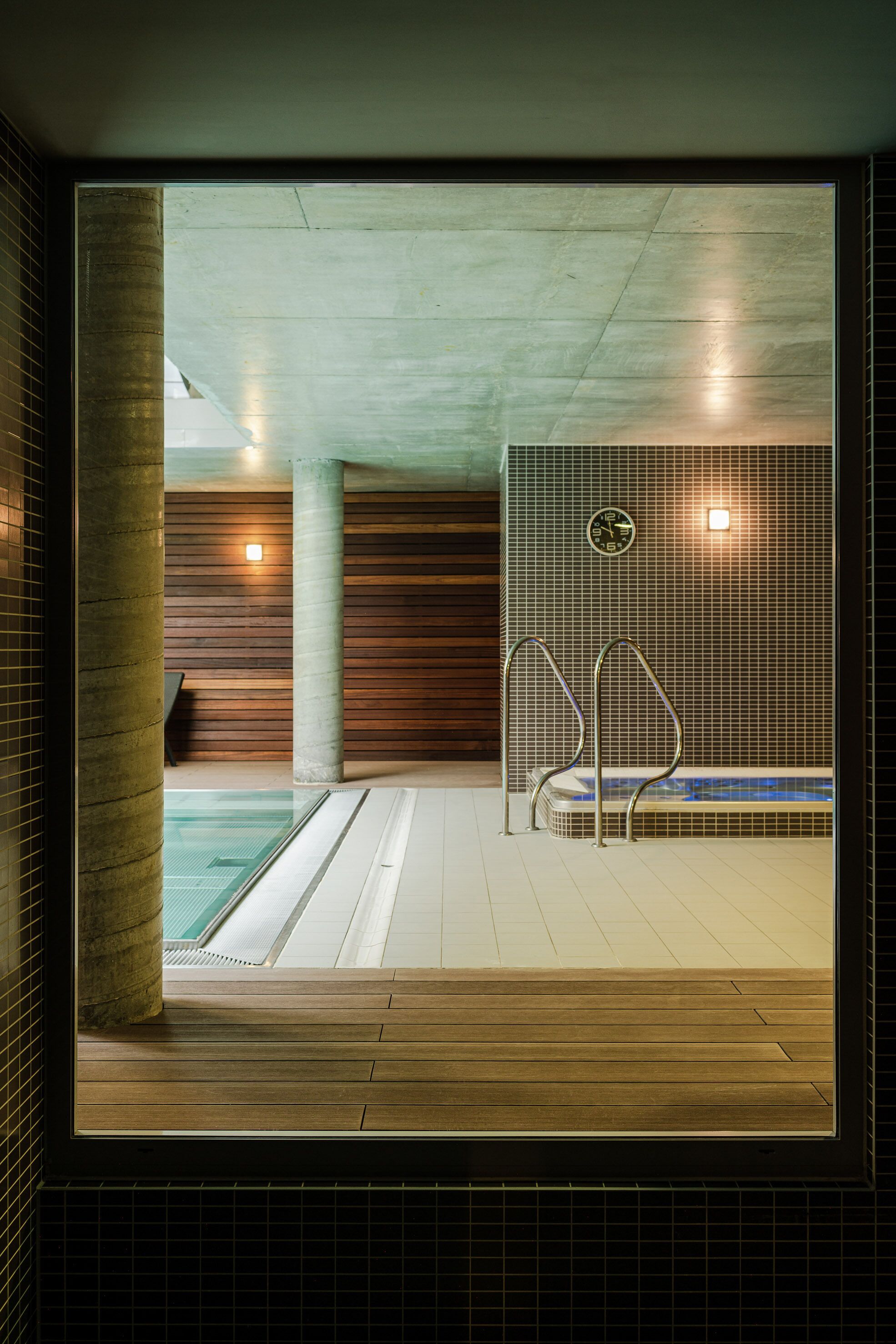 Un área de piscina interior moderna con un deck de madera, una bañera de hidromasaje y un reloj visible en la pared.