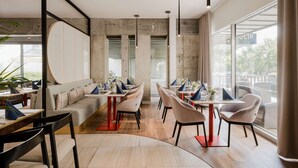 Un comedor moderno con una combinación de banquetas y mesas, que presenta una zona de estar central con un sofá y sillas, y grandes ventanales con vistas al exterior.