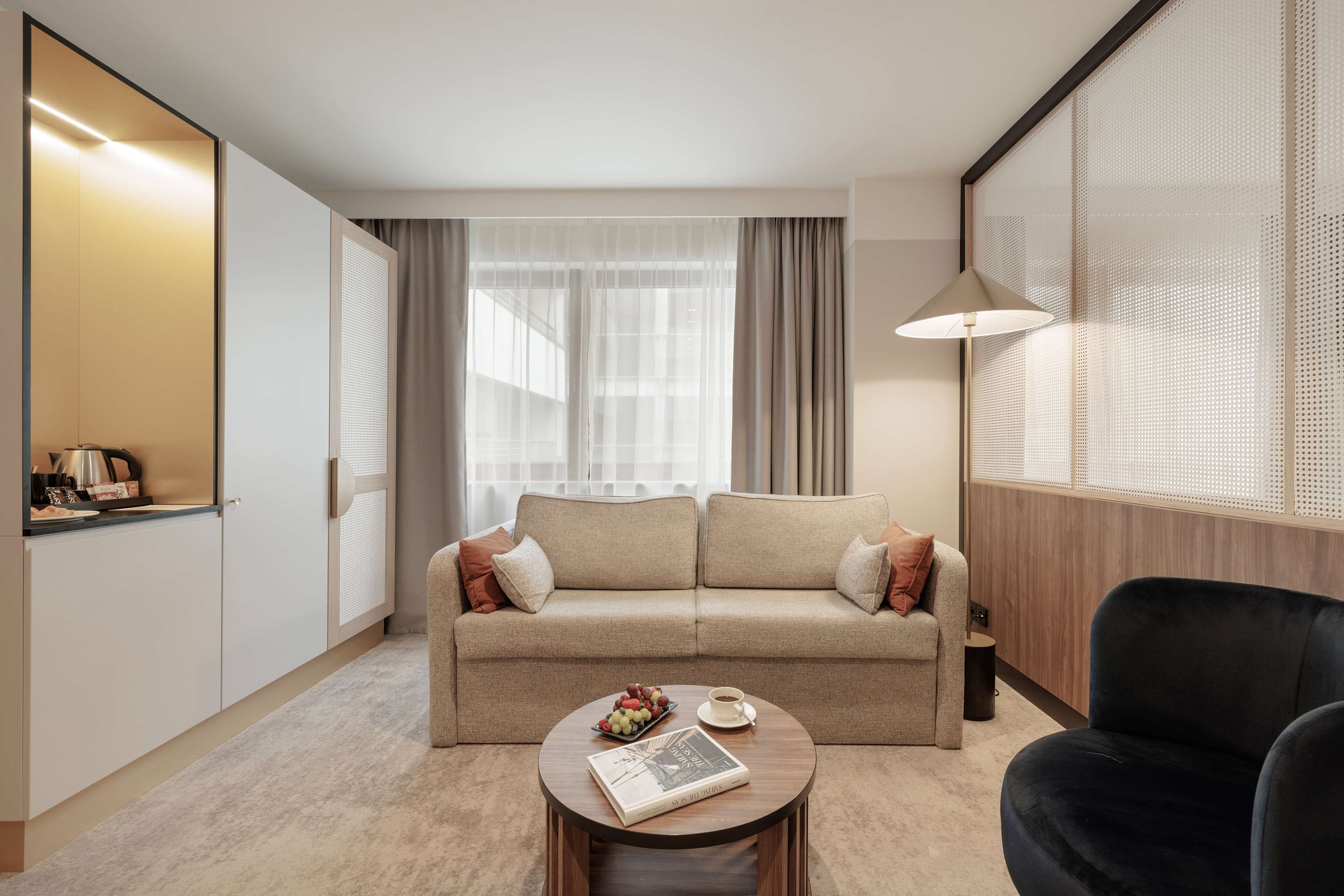 A modern living room with a beige sofa, a round coffee table with a book and fruit, a black armchair, and a large window with sheer curtains.
