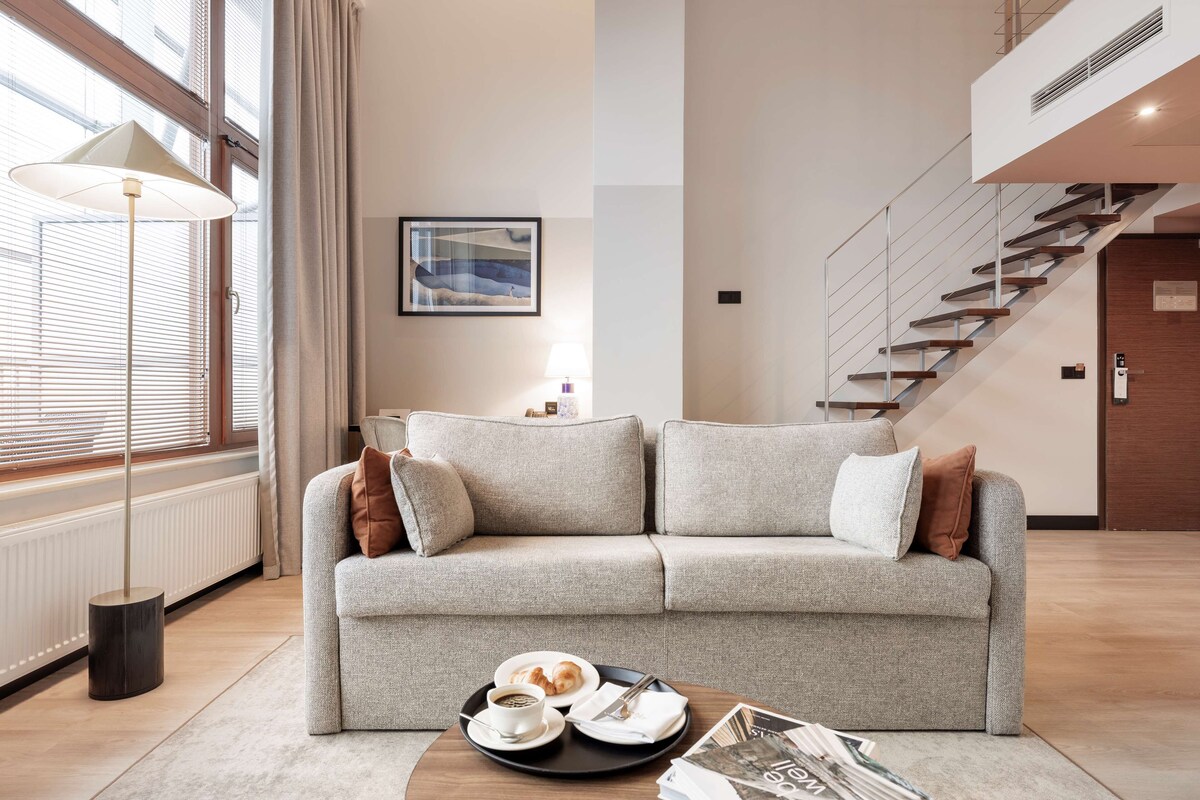Un salon moderne avec un canapé gris, une table basse sur laquelle est posée une assiette de pâtisseries, et un escalier en arrière-plan.