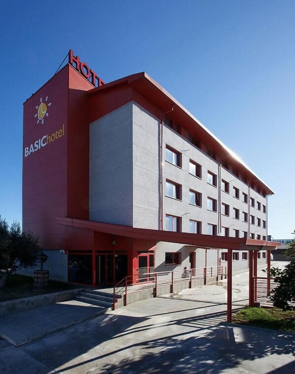 A multi-story hotel with a red and grey facade, a visible logo, and a staircase leading to the entrance. - Hotel Basic (Olerdola)