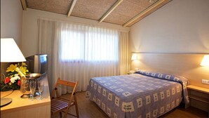Habitación de hotel con cama, escritorio con lámpara, silla, televisor y ventana con cortinas.