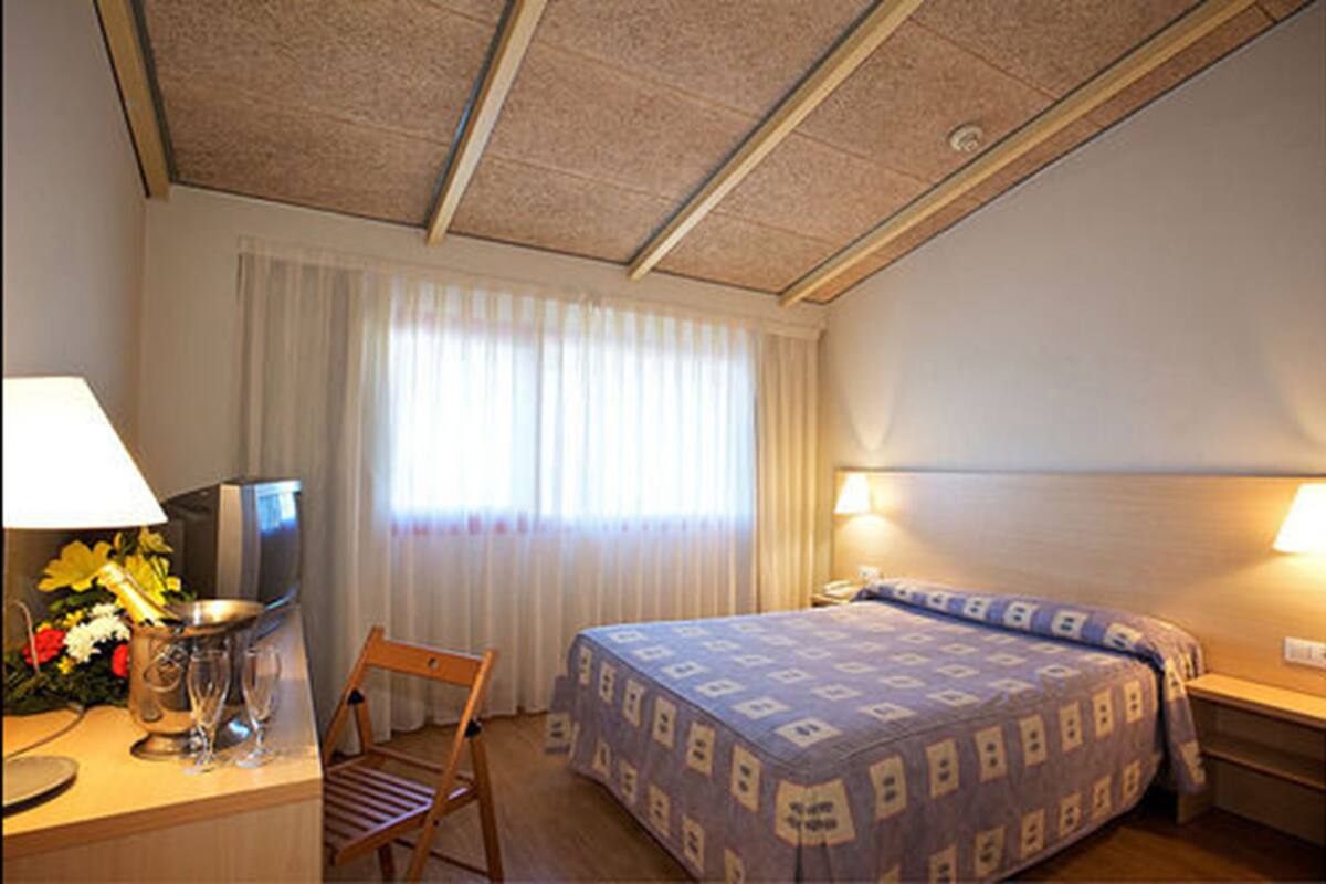 A hotel room with a bed, a desk with a lamp, a chair, a TV, and a window with curtains.