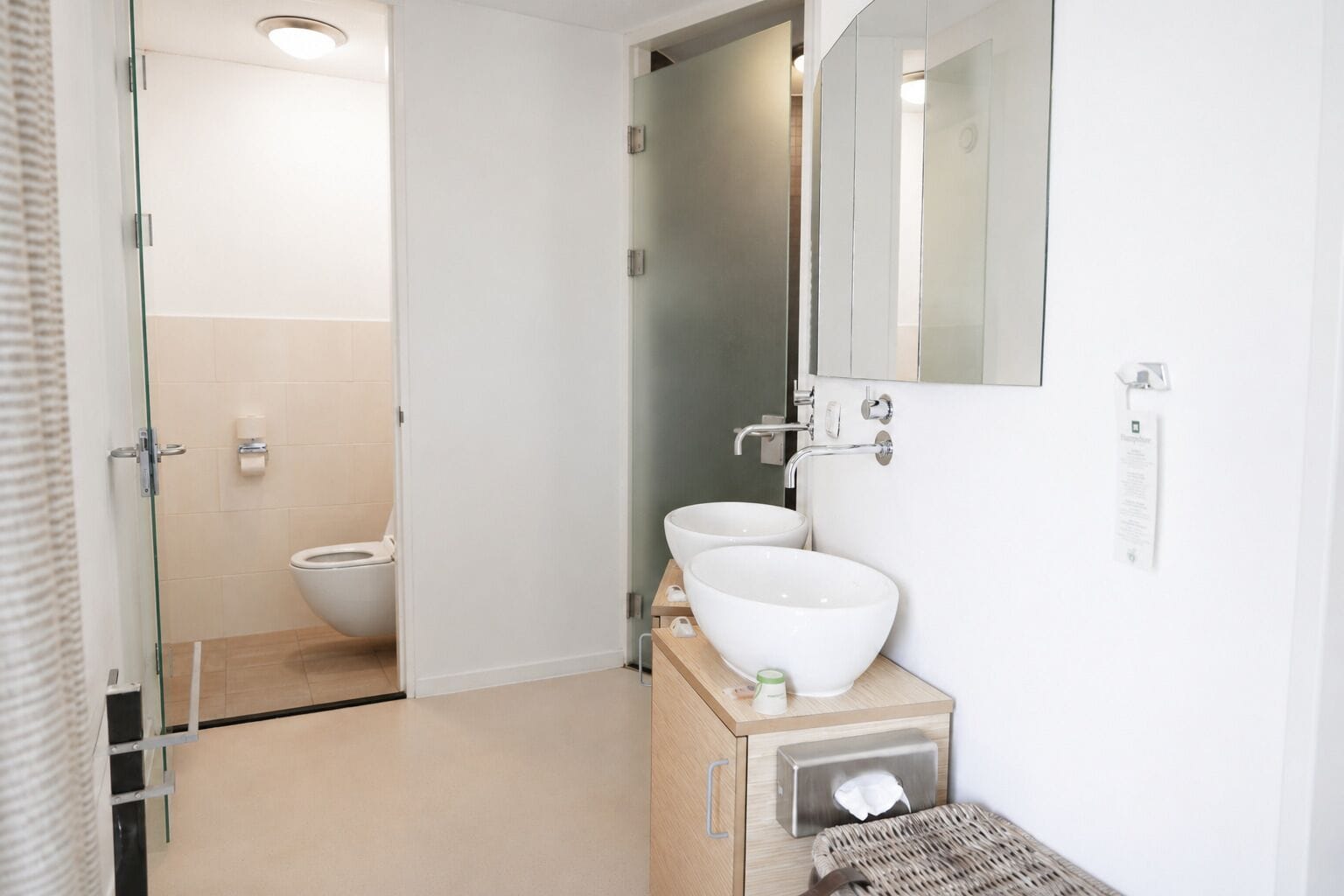 Een moderne badkamer met toilet, twee wasbakken, een spiegel en een handdoekdispenser.