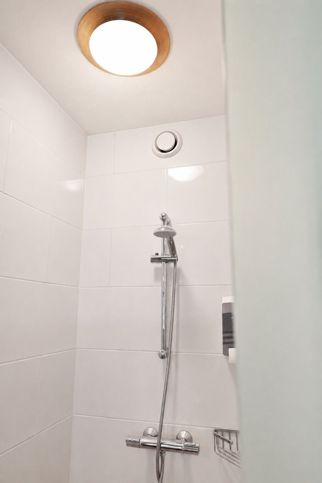 A shower with a handheld showerhead and a wall-mounted faucet, a round light fixture on the ceiling, and a corner-mounted soap dispenser.