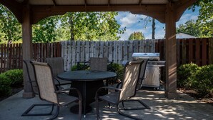 A covered patio with a round table and four chairs, a grill, and a wooden fence in the background.
