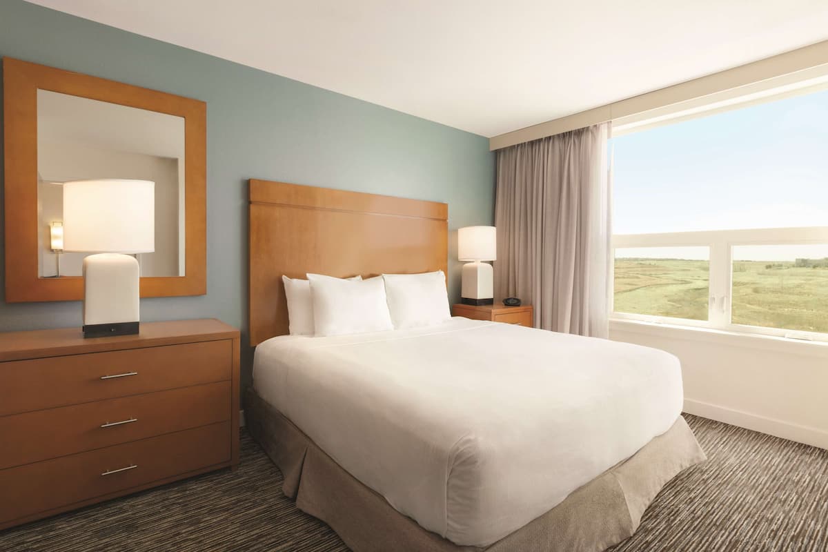 A hotel room with a large bed, a wooden headboard, a dresser with drawers, two bedside lamps, and a large window with a view of the countryside.