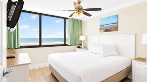 A hotel room with a bed, a TV, a ceiling fan, a nightstand, a lamp, a painting, and a view of the beach. - Sands Beach Club (Myrtle Beach)