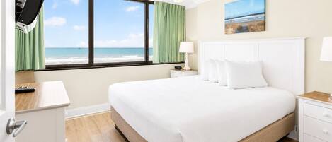 A hotel room with a bed, a TV, a ceiling fan, a nightstand, a lamp, a painting, and a view of the beach.