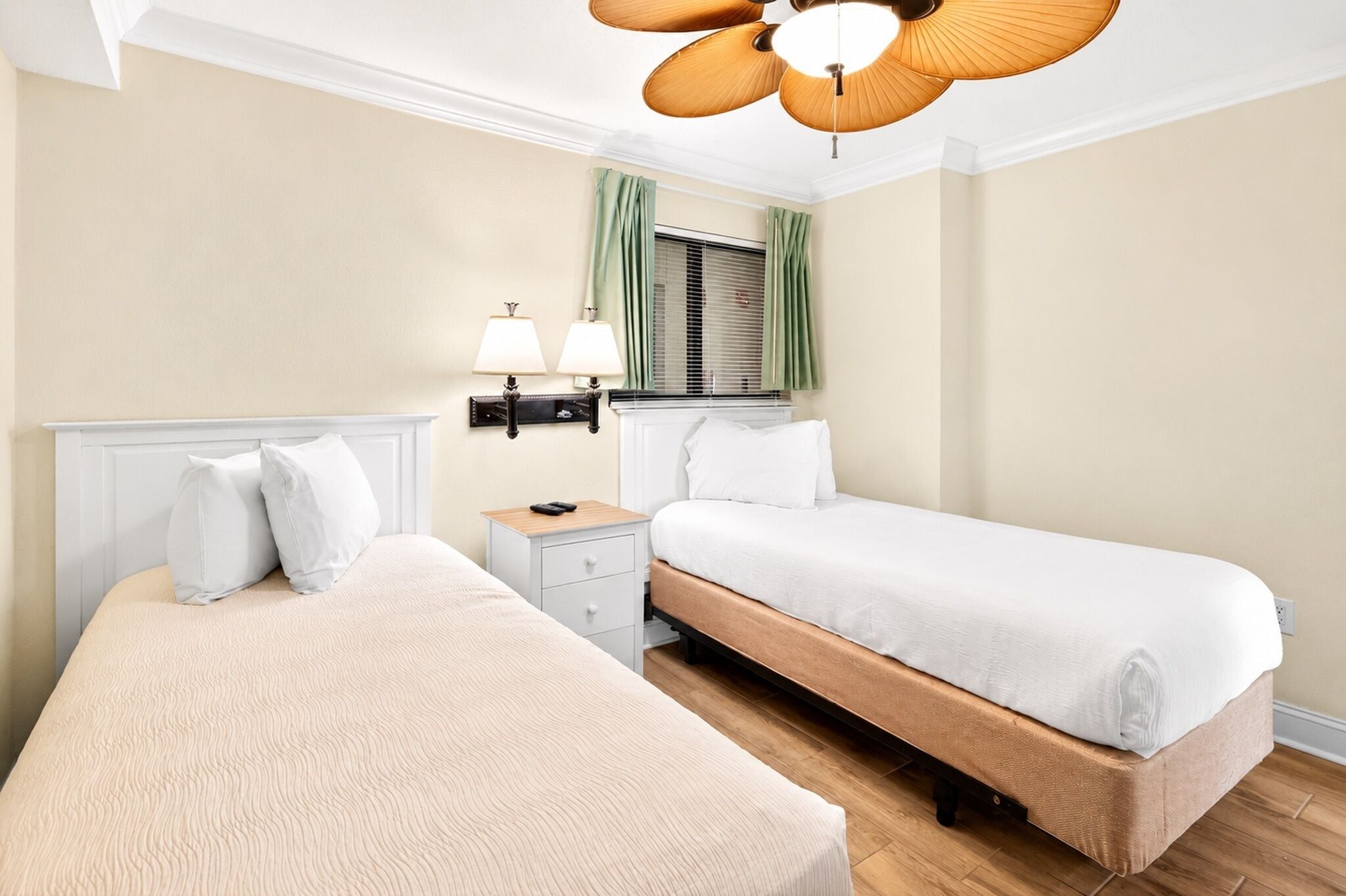 Ein Hotelzimmer mit zwei Betten, einem Nachttisch mit Lampen, einem Fenster mit Vorhängen und einem Deckenventilator.