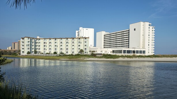 A hotel with multiple floors, a large building with a flat roof, and a body of water in the foreground. - Sands Beach Club (Myrtle Beach)