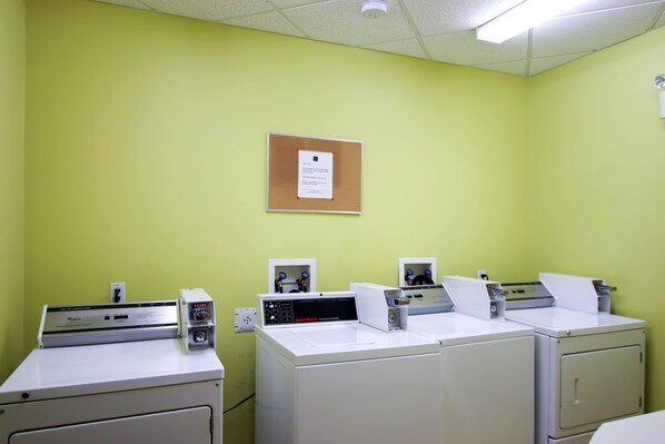 A laundry facility with multiple washing machines and dryers, a bulletin board with a notice, and a fluorescent ceiling light.