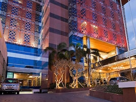A modern building with a glass facade, a red patterned facade, and a well-lit entrance area with palm trees.