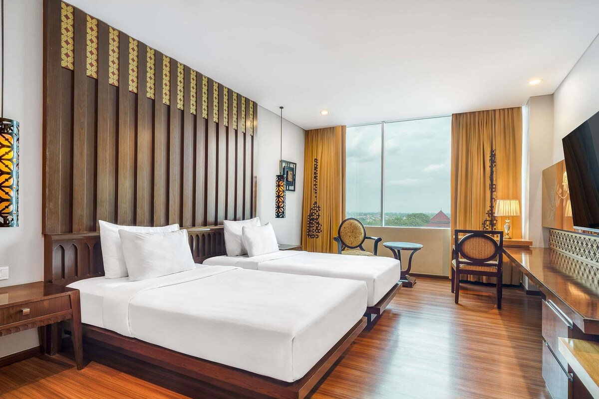 A hotel room with two beds, a desk, a chair, a small table, and a large window offering a city view.