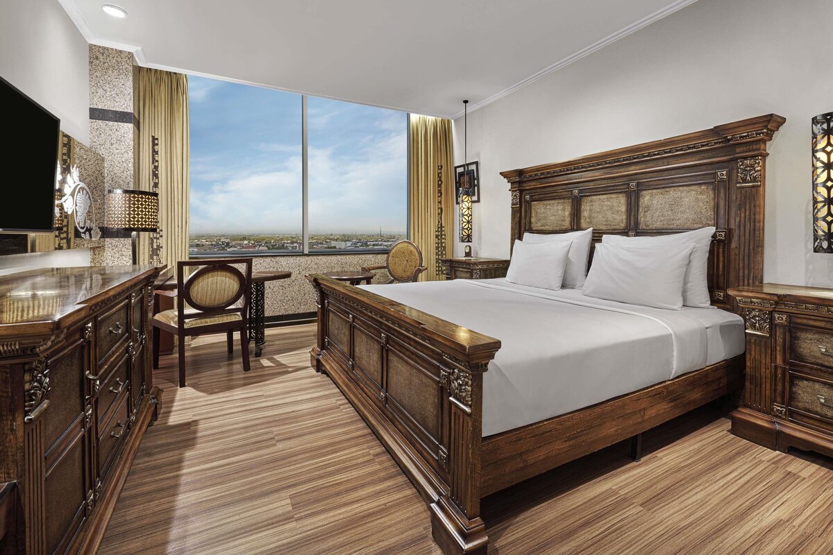 A spacious bedroom with a large bed, wooden furniture, and a city view through large windows.