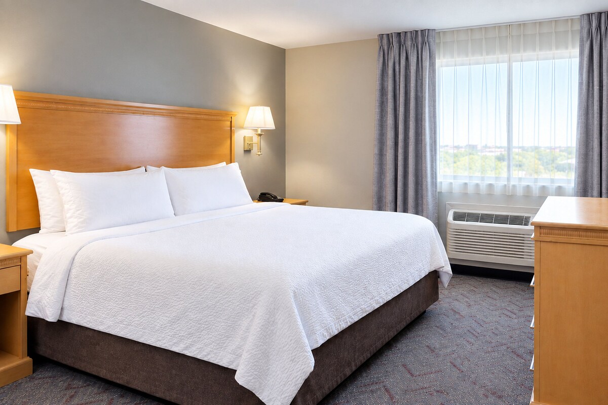 A hotel room with a large bed, a bedside table, a lamp, a TV, and a window with curtains.