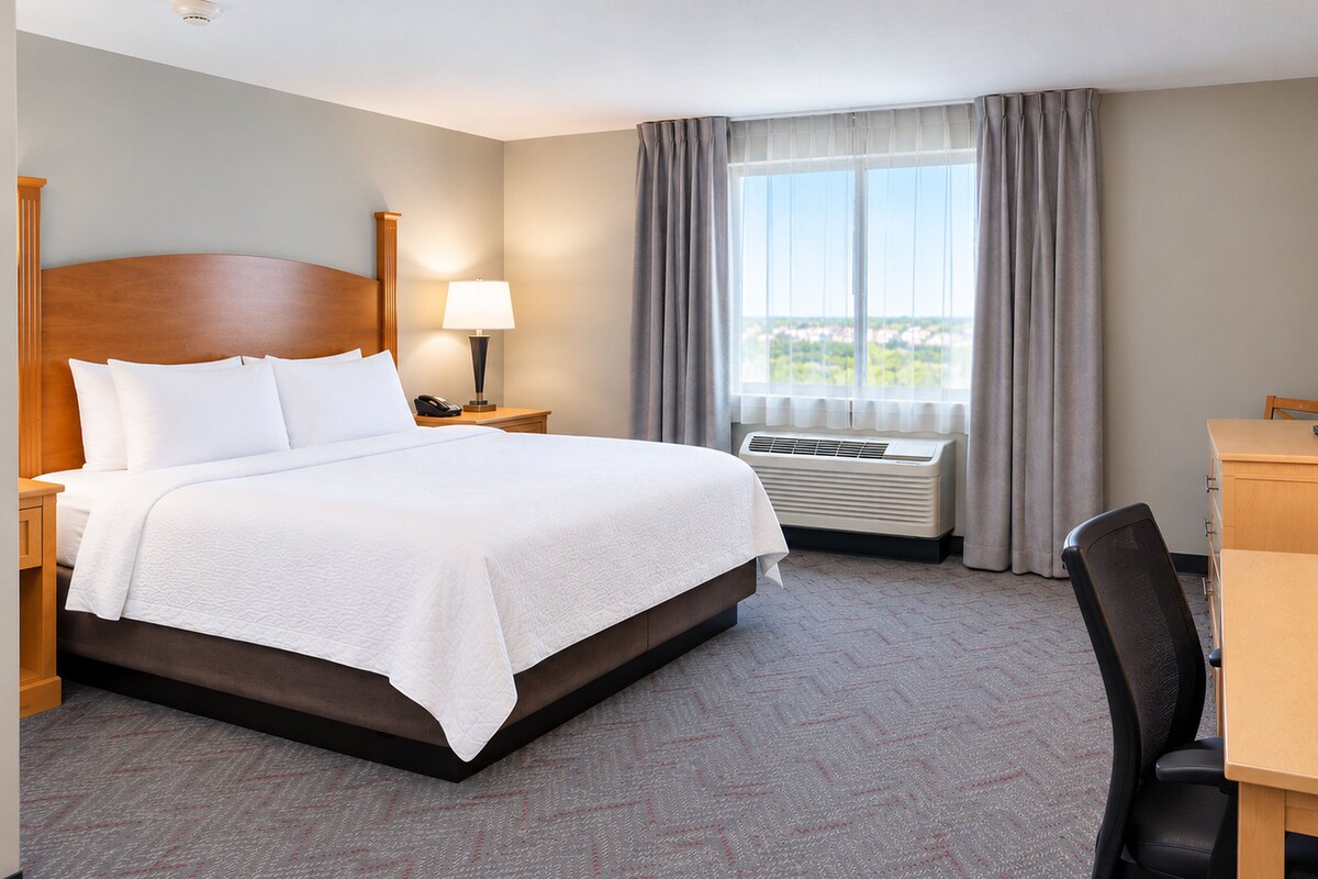 A hotel room with a bed, bedside table, lamp, TV, desk, chair, and a window with curtains.