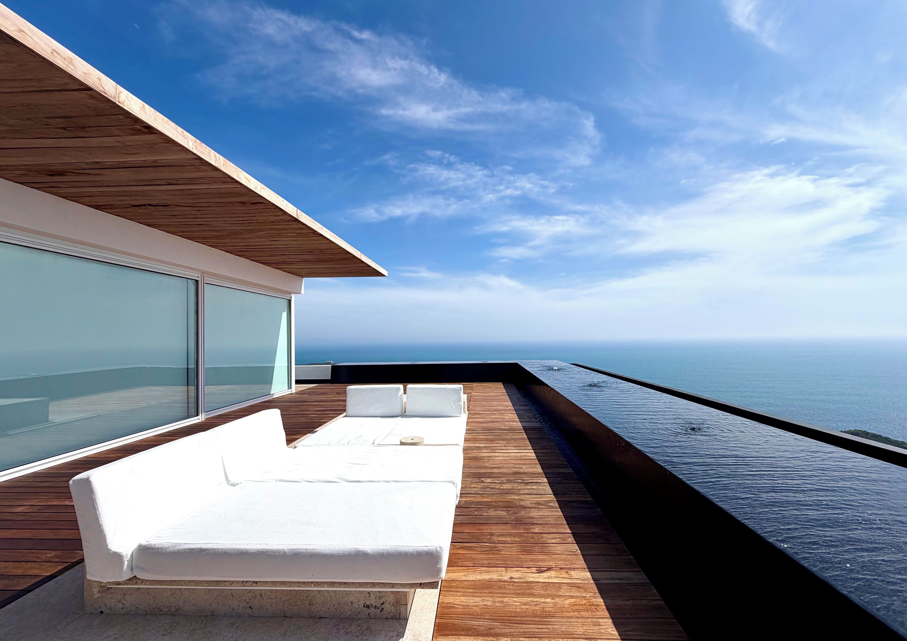 Une terrasse extérieure moderne avec un plancher en bois, des sièges blancs rembourrés et un mur de verre donnant sur la mer.