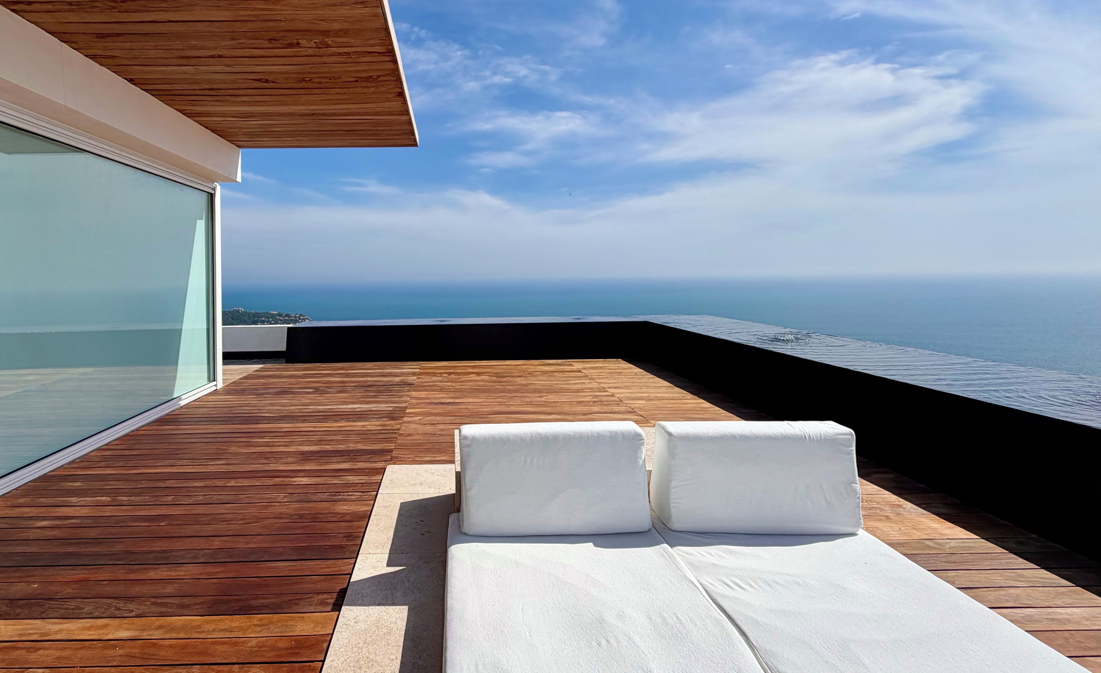 Une terrasse en bois avec un espace salon agrémenté de coussins blancs, donnant sur une étendue d’eau.