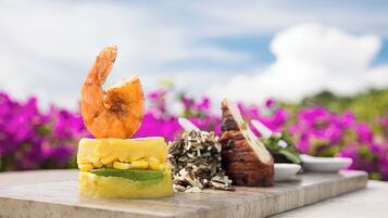 A variety of food items on a wooden board, including a shrimp, a slice of bread, and a stack of corn and avocado.