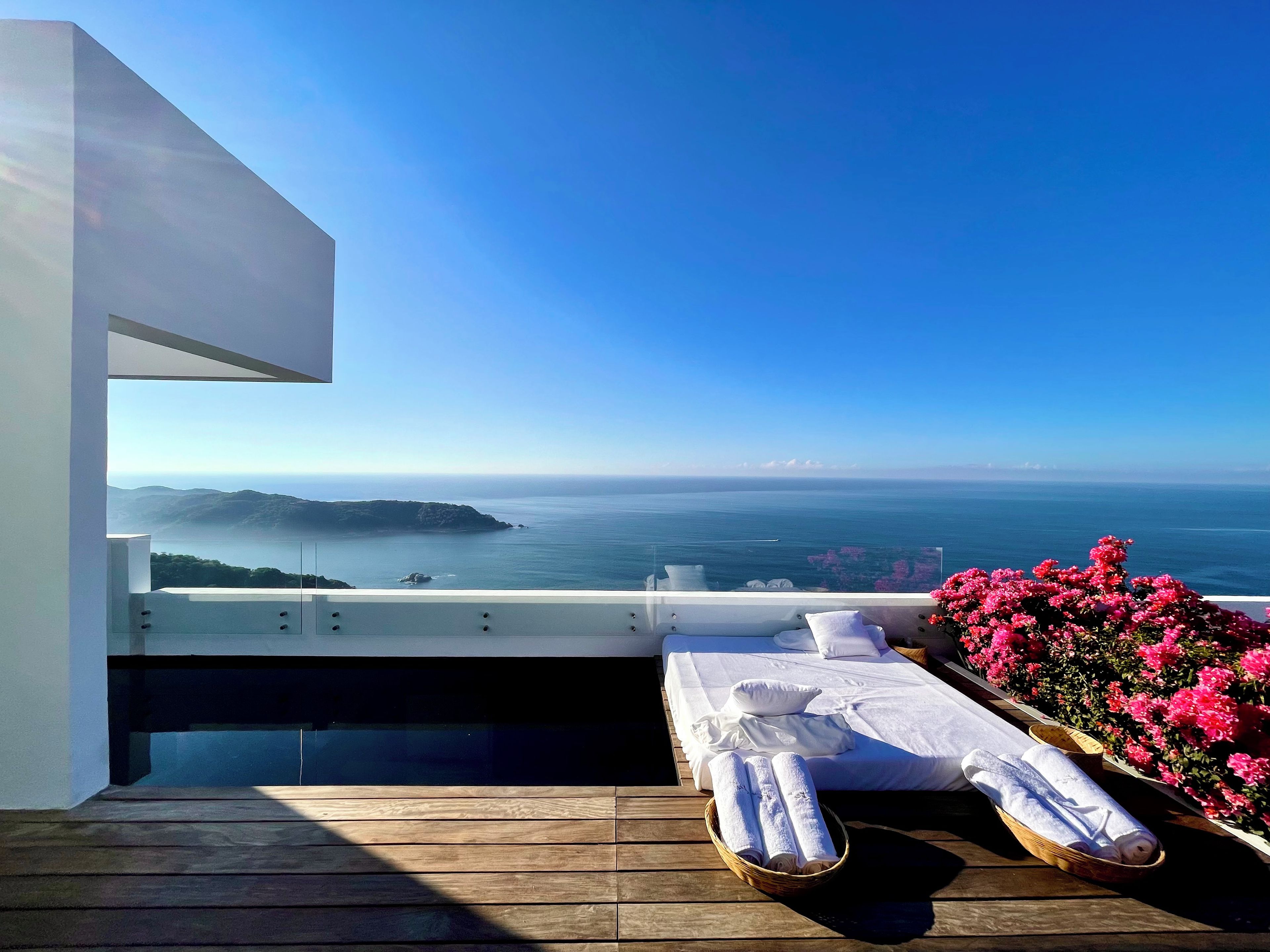 A balcony with a wooden floor, a bed with white linens, and a view of the sea.