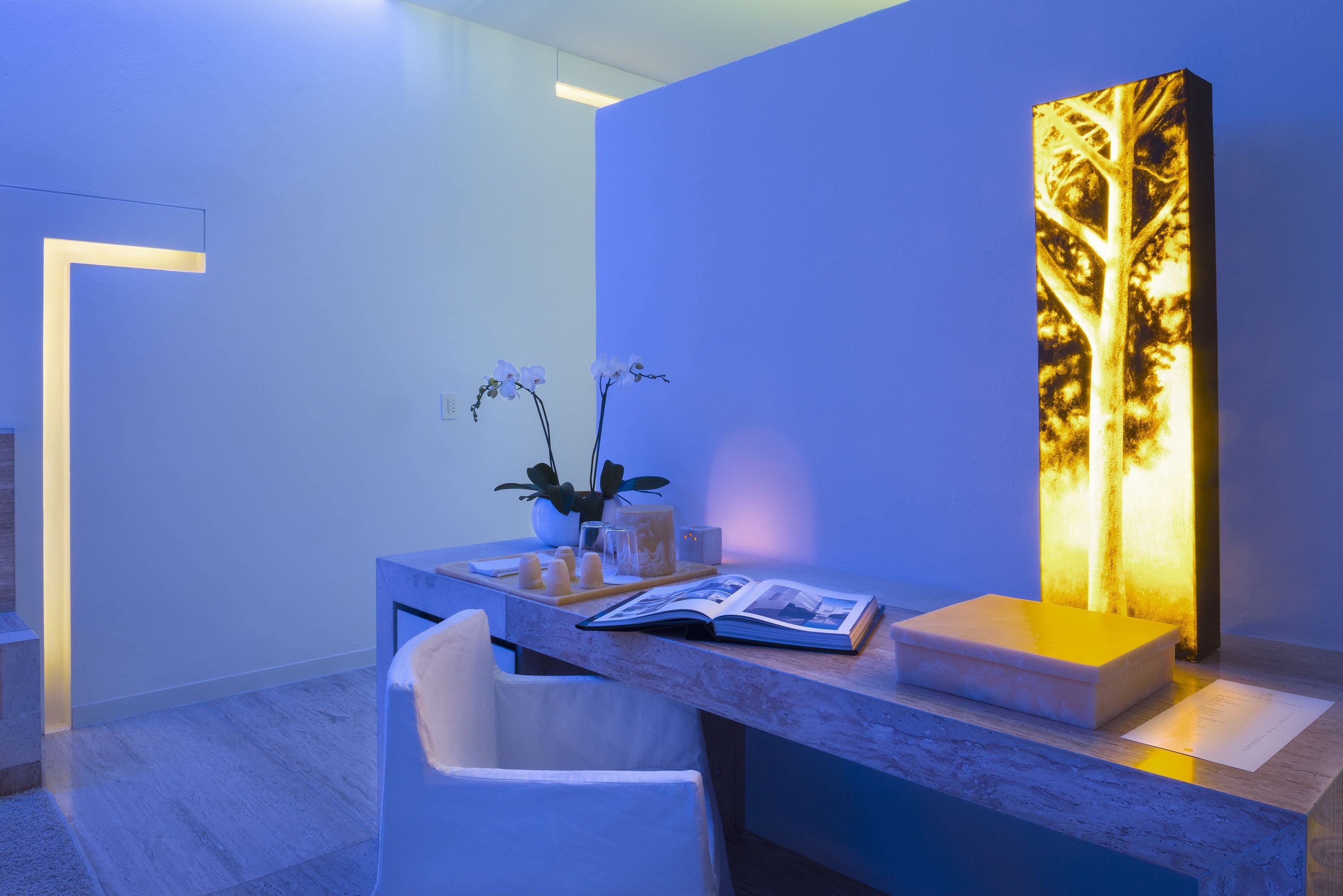 A modern reception desk with a marble top, a white chair, a vase with orchids, and a lit vertical panel.