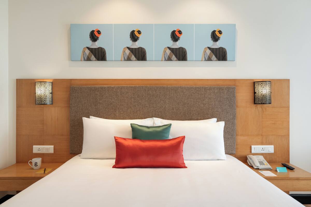 Habitación de hotel con cama, almohadas, mesita de noche, teléfono, control remoto y una obra de arte en la pared.