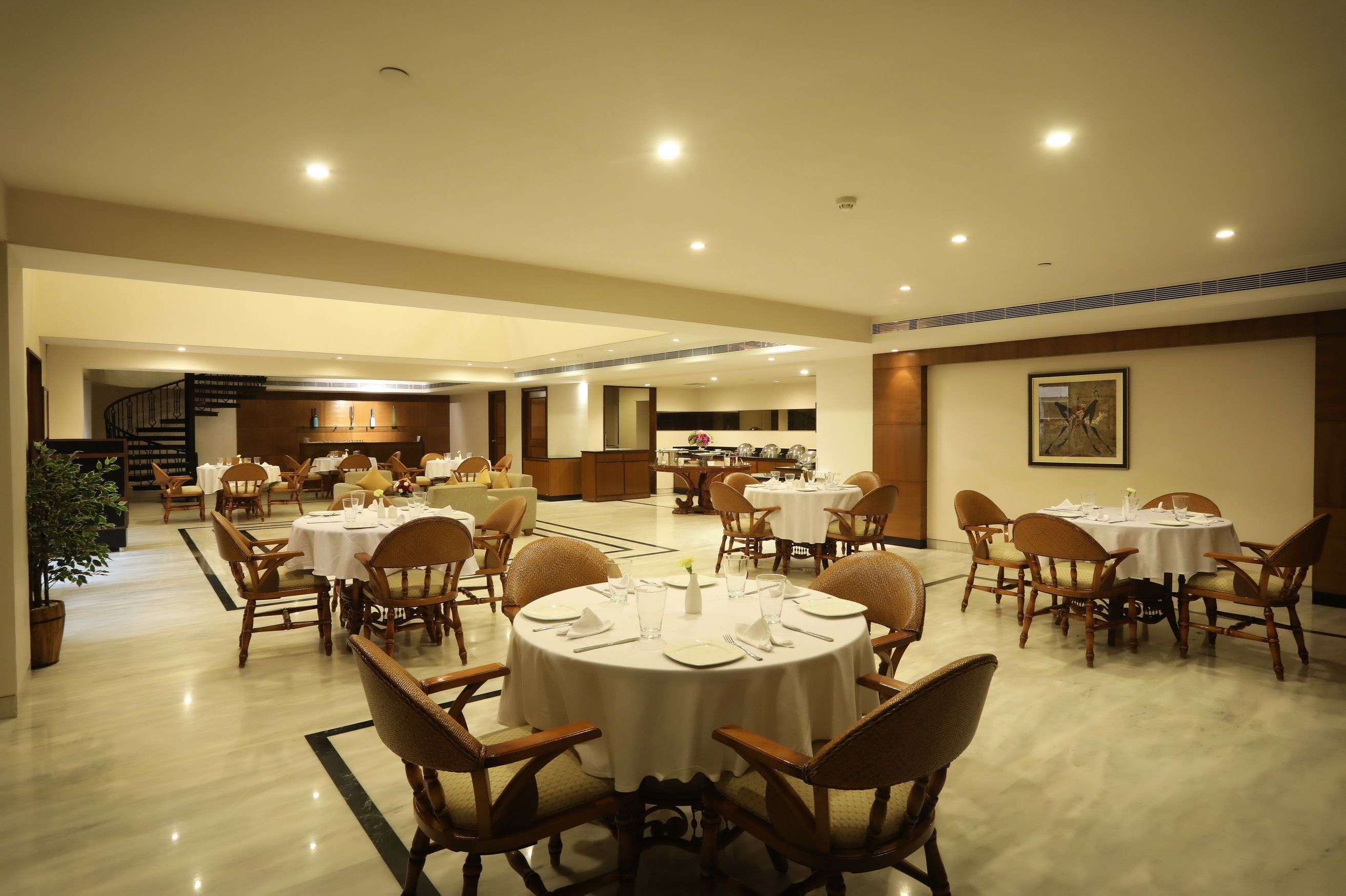 A spacious dining area with round tables and chairs, a staircase in the background, and a kitchen area visible through an open door.
