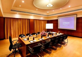 A conference room with a long table, chairs, a projector screen displaying "HITACHI", and a floral arrangement.