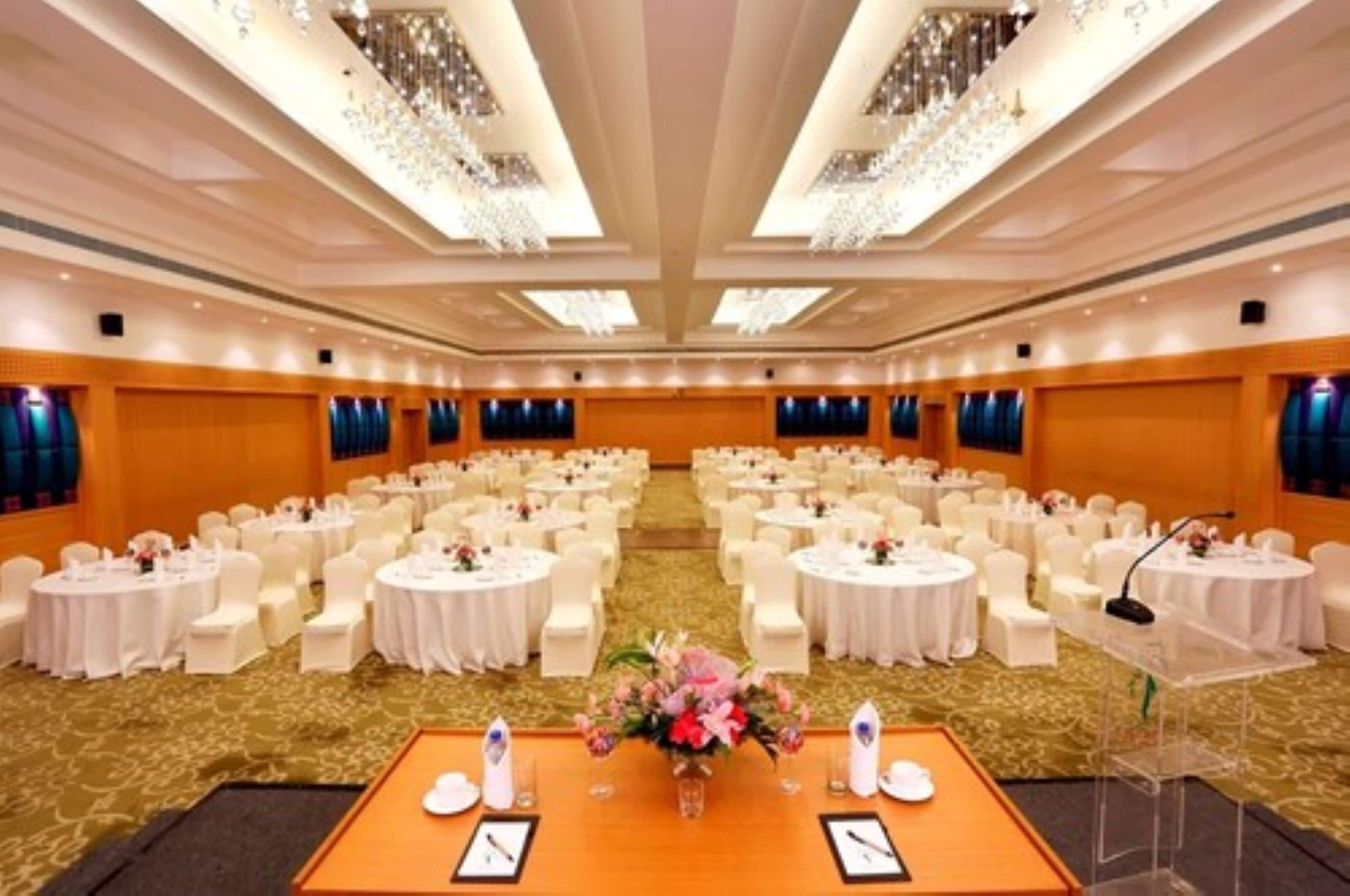 Un amplio salón de conferencias con mesas redondas y sillas blancas, un atril y una decoración floral.