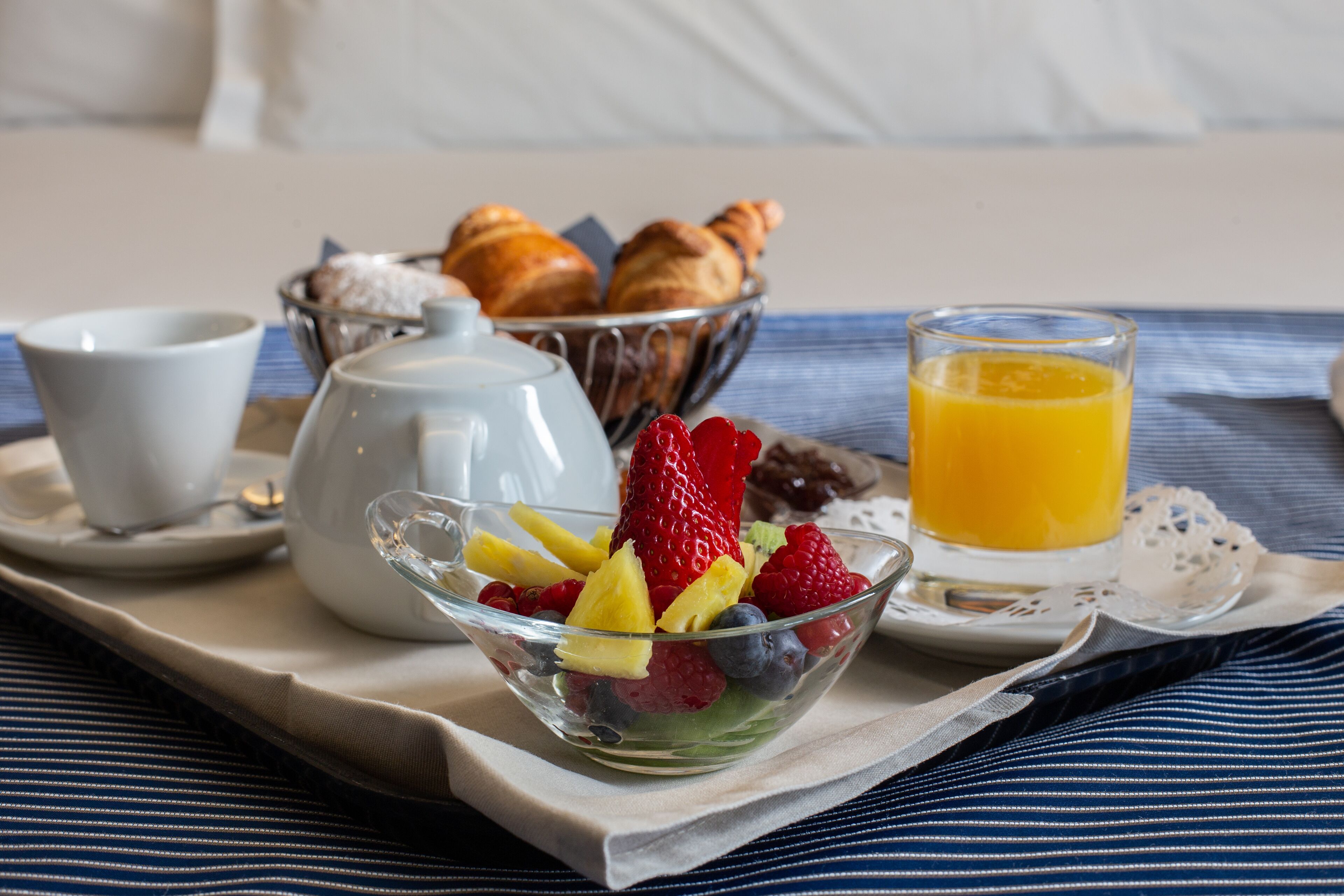 Una bandeja de desayuno con croissants, una tetera, un cuenco de fruta, una taza y un vaso de zumo de naranja.