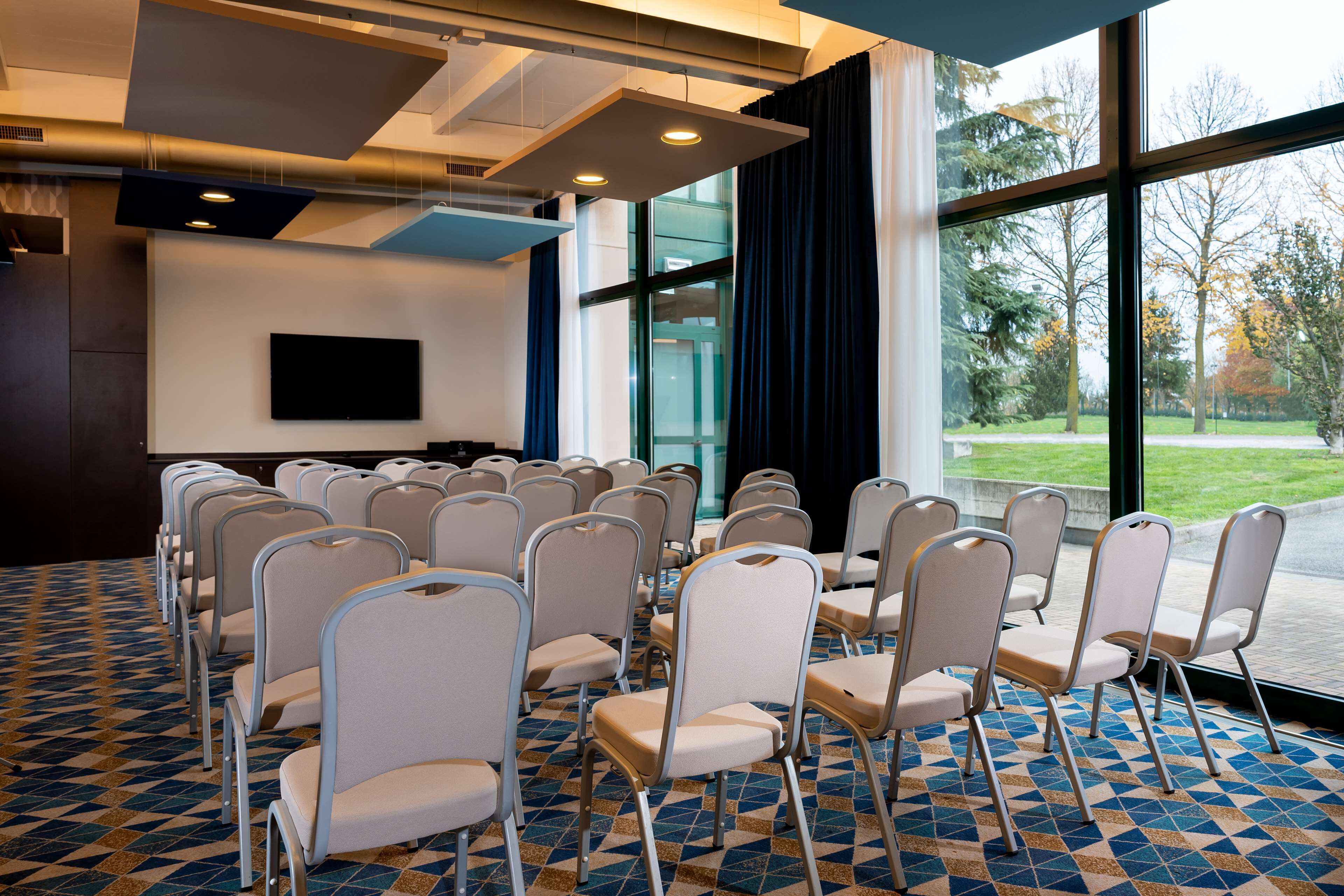 Un salón de conferencias con filas de sillas, un televisor de pantalla plana en la pared y grandes ventanales con vistas a árboles.