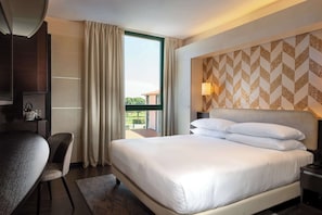 Habitación de hotel con una cama grande, un escritorio y una silla. Se ve una ventana con cortinas, una pared con un diseño geométrico y una mesita de noche con una lámpara.