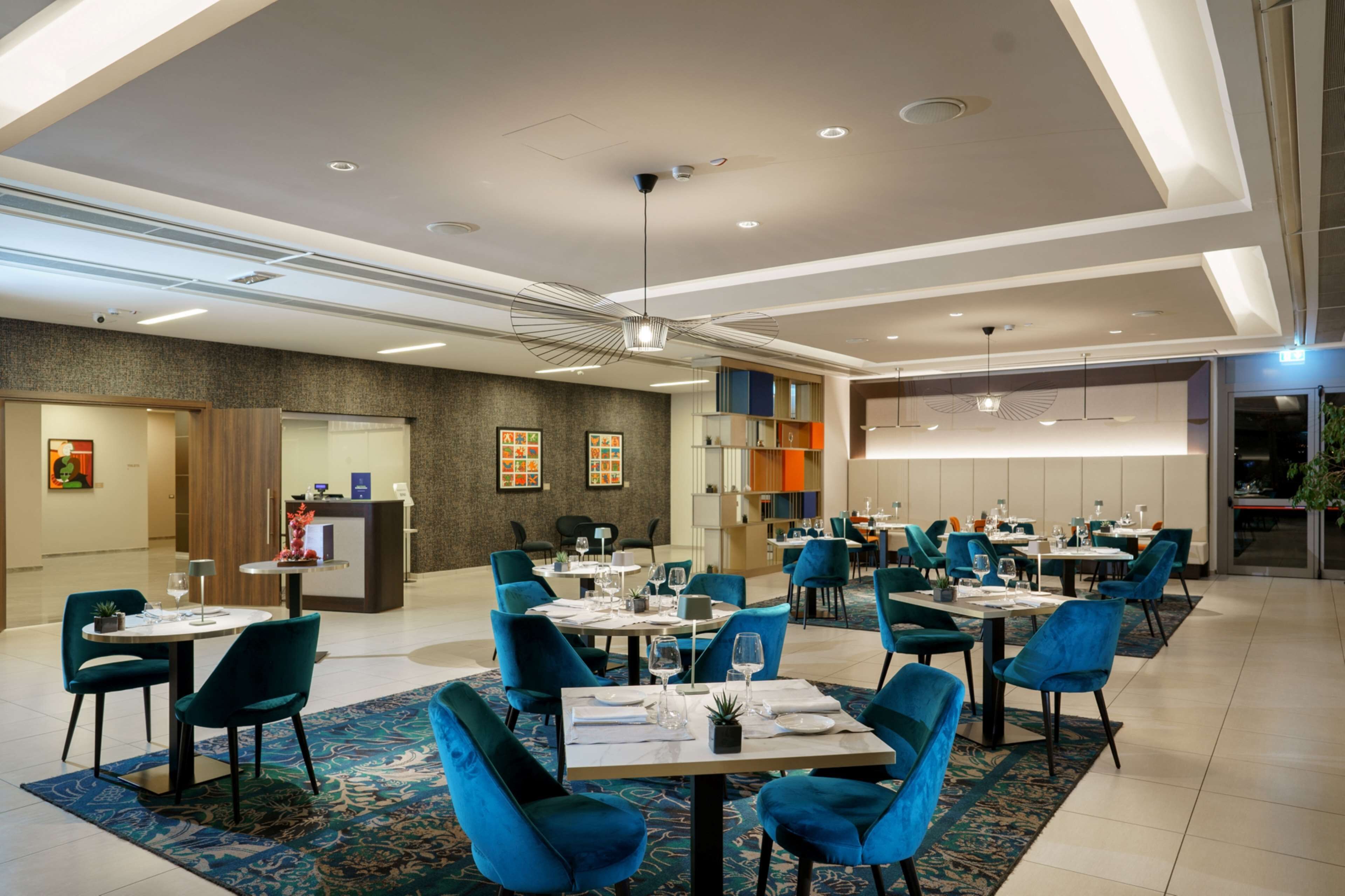 Un área de comedor moderna con sillas y mesas azules, una recepción y un ventanal grande.