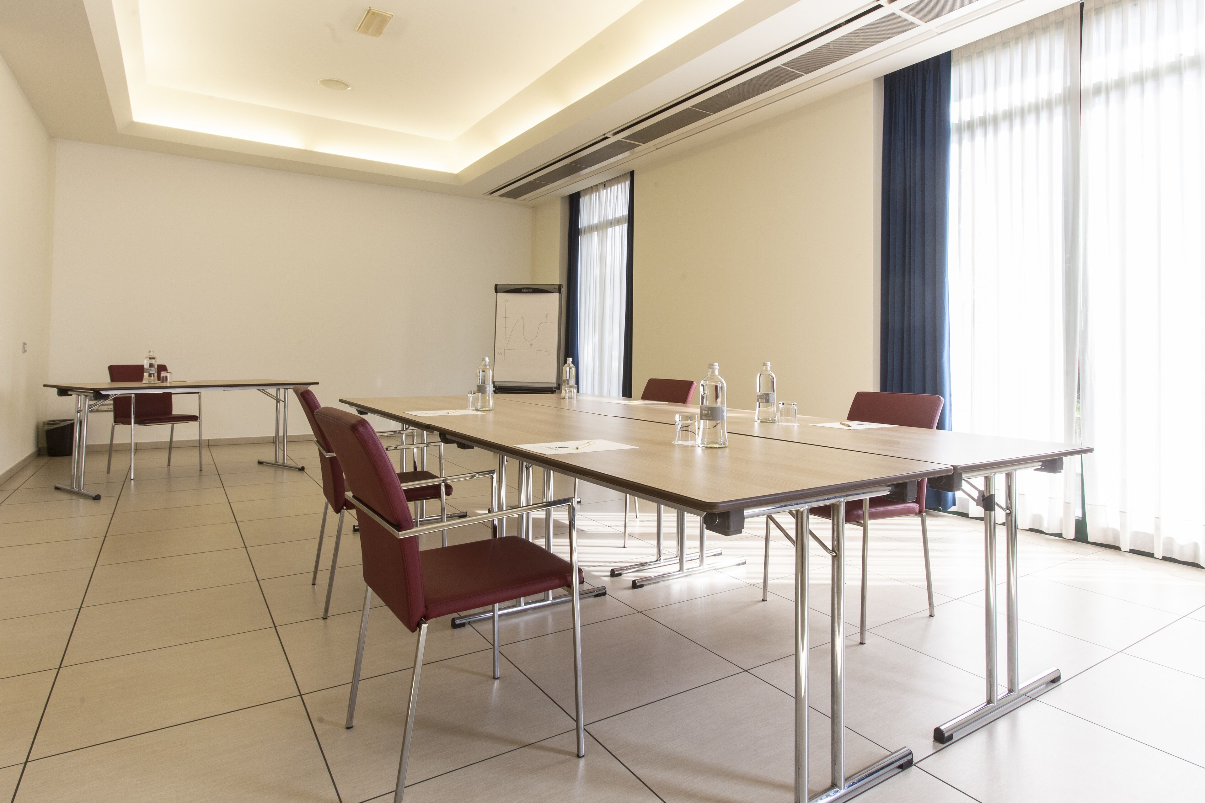 A conference room with a long table, red chairs, a whiteboard, and large windows with curtains.