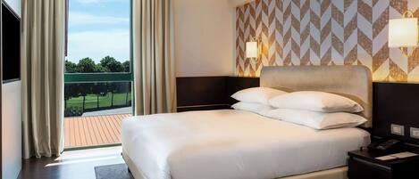 A hotel room with a large bed, a view of the outdoors, a patterned wall, and a nightstand with a lamp.