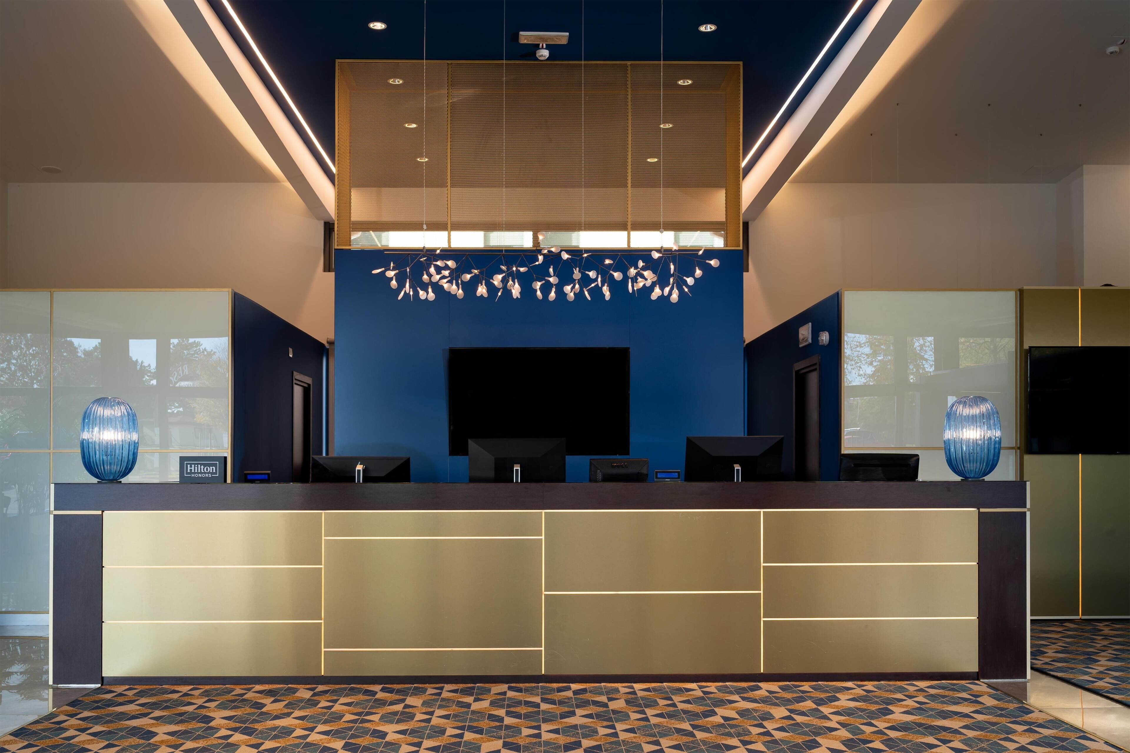 A modern hotel reception area with a blue accent wall, a large flat-screen TV, and a decorative ceiling light fixture.