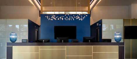 A modern hotel reception area with a blue accent wall, a large flat-screen TV, and a decorative ceiling light fixture.