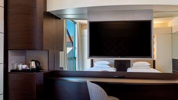 A modern hotel room with a large flat-screen TV, a seating area with a chair, a coffee station, and a view of the outdoors.