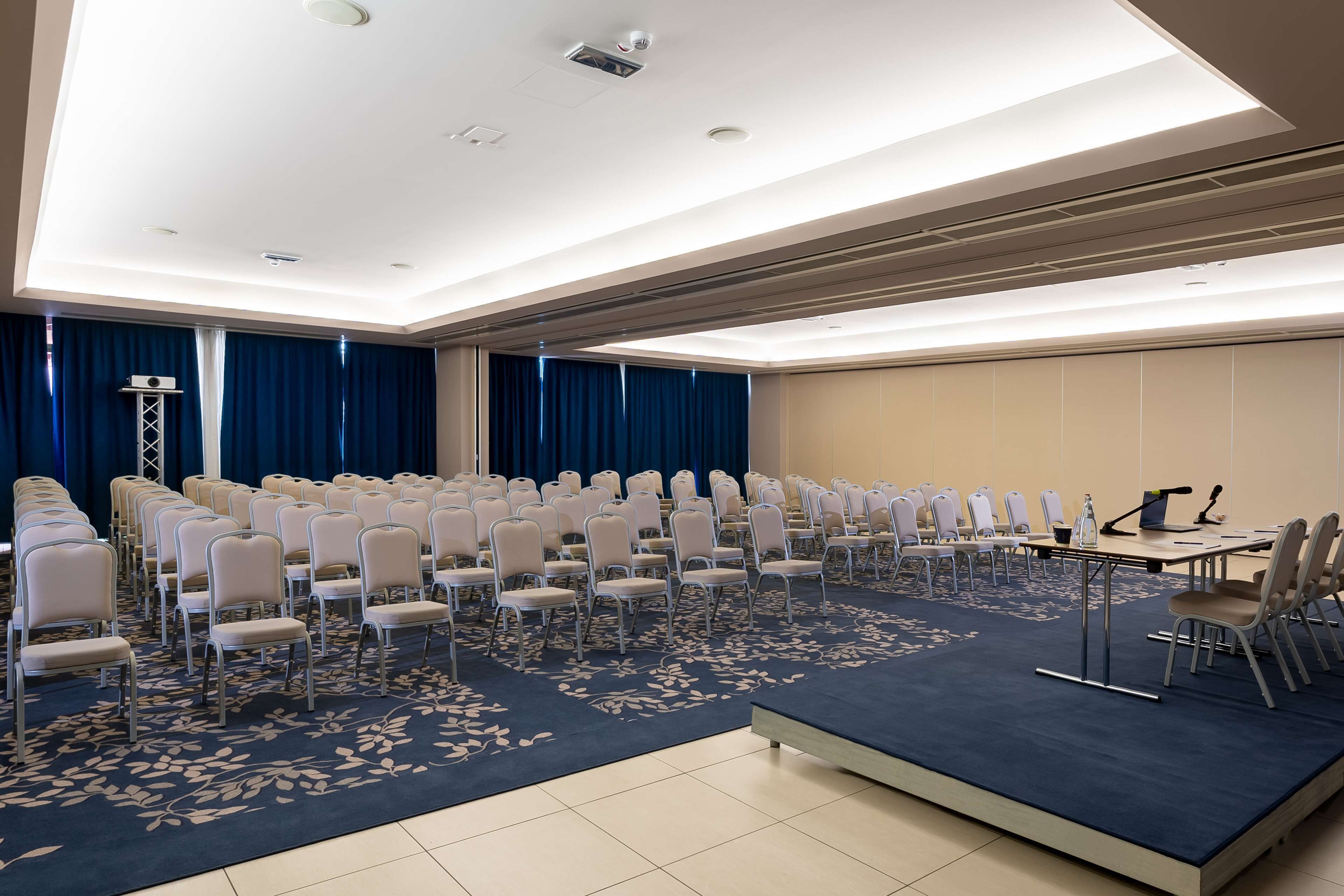 Une salle de conférence avec des rangées de chaises, une scène et un sol recouvert de moquette.