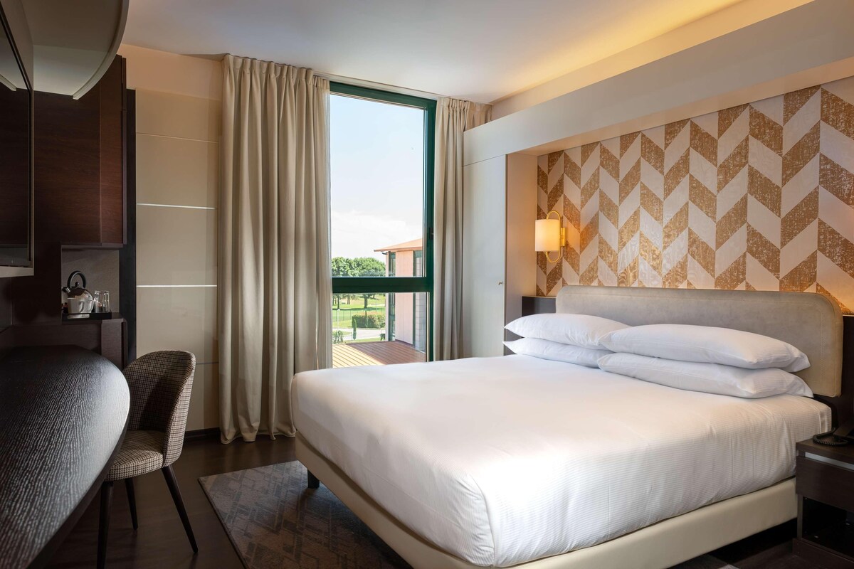 A hotel room with a large bed, a desk, and a chair. There is a window with curtains, a wall with a geometric pattern, and a nightstand with a lamp.