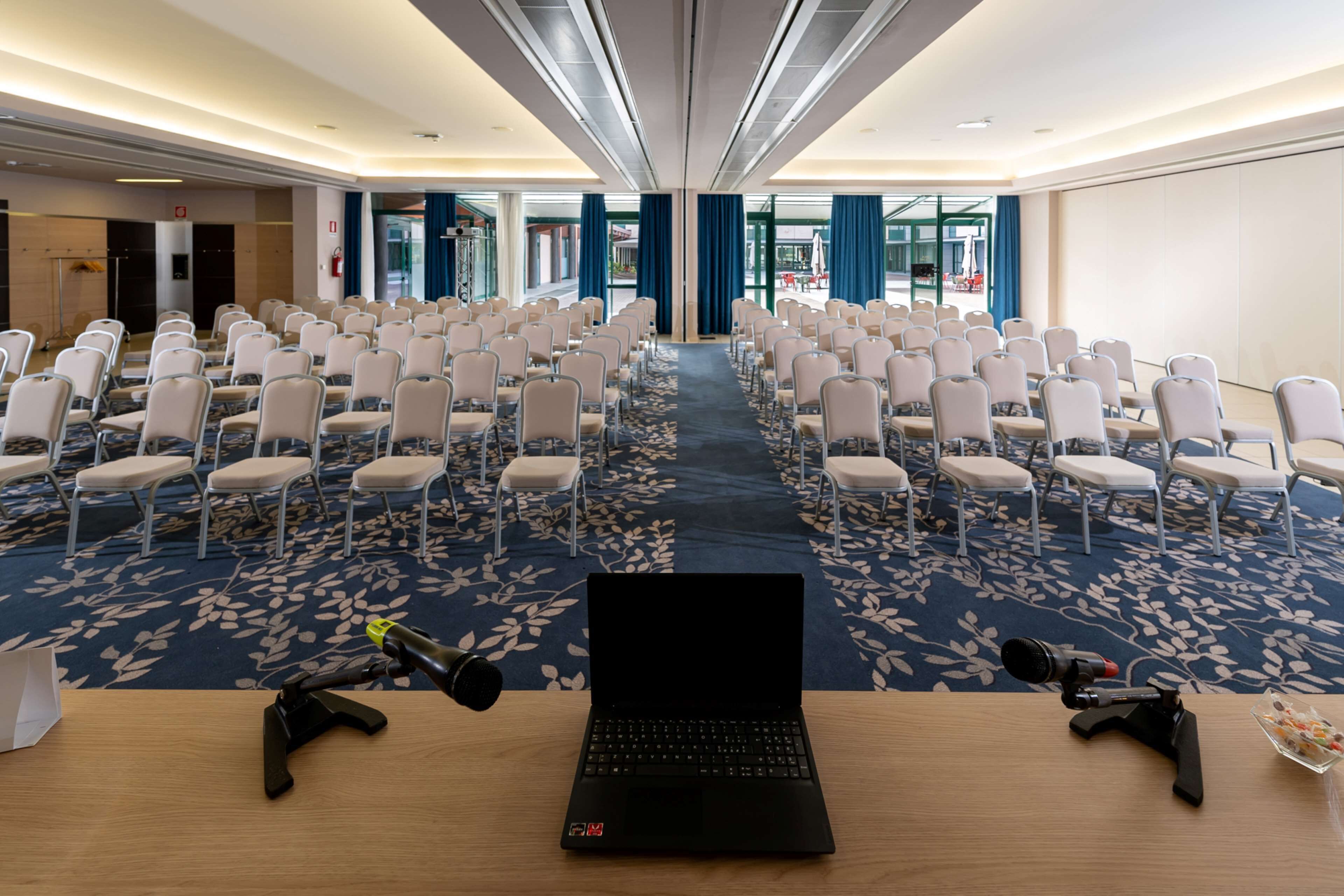 Un salón de conferencias con filas de sillas, un ordenador portátil sobre una mesa y una cámara sobre un trípode.