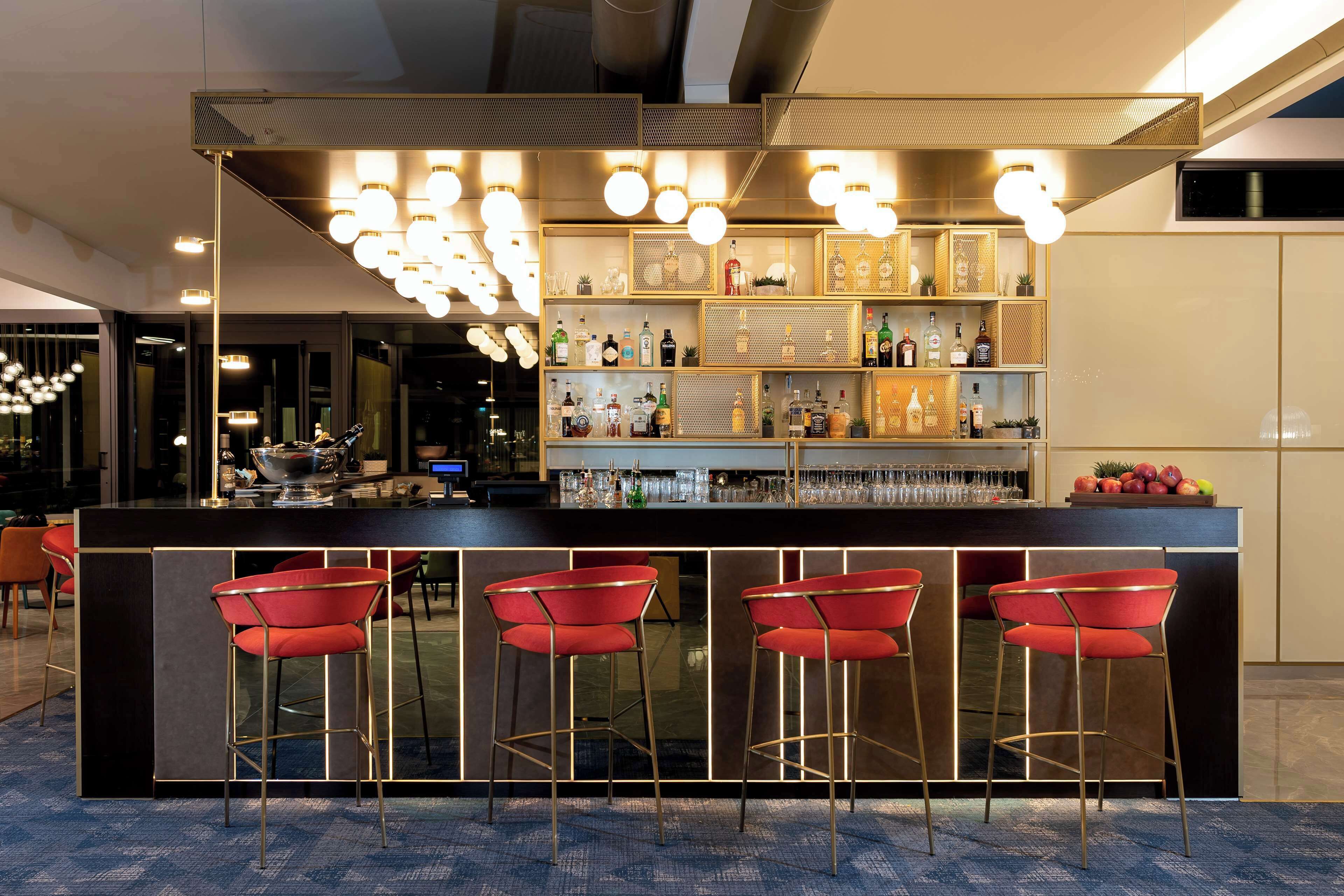 A modern bar area with a dark counter, red bar stools, and a well-lit ceiling with multiple hanging lights.